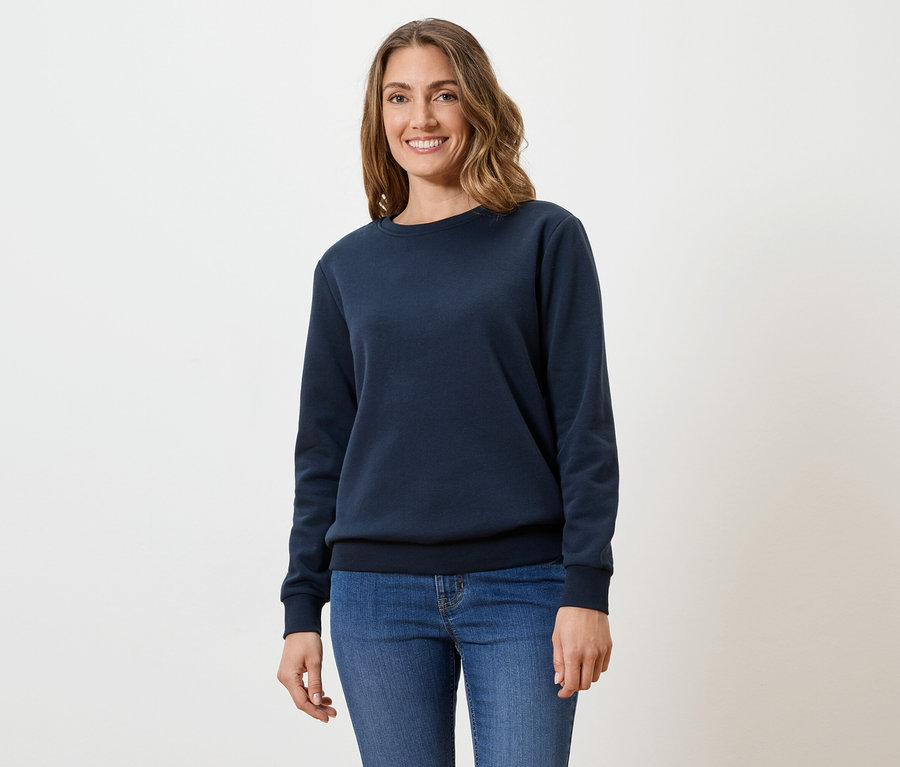 Une femme aux cheveux bruns ondulés porte un pull bleu marine et un jean sur un fond blanc.