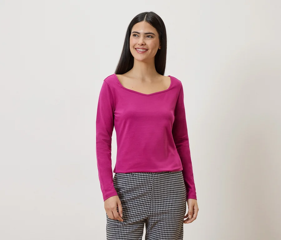 Une femme aux longs cheveux foncés porte un t-shirt rose à manches longues et un pantalon à carreaux noirs et blancs sur un fond blanc.