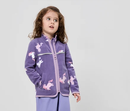 Une fillette aux cheveux bruns bouclés porte une veste en polaire violette à motifs de licornes et un pantalon violet sur un fond neutre.