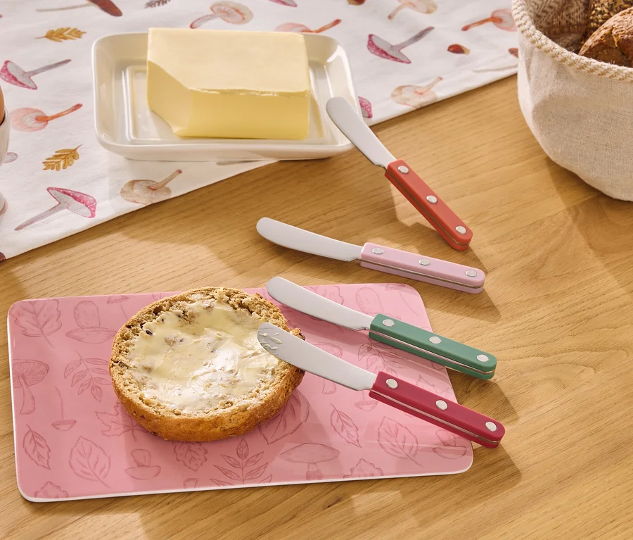 Sur une table en bois sont posés un œuf dans un coquetier, du beurre, un pain beurré et quatre couteaux à beurre de couleurs rouge, rose, verte et rouge.