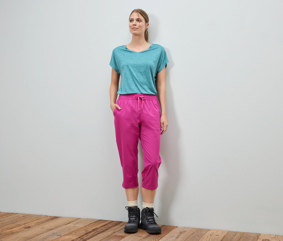 Une femme se tient devant un mur et porte un t-shirt bleu, un pantalon rose et des bottes noires.
