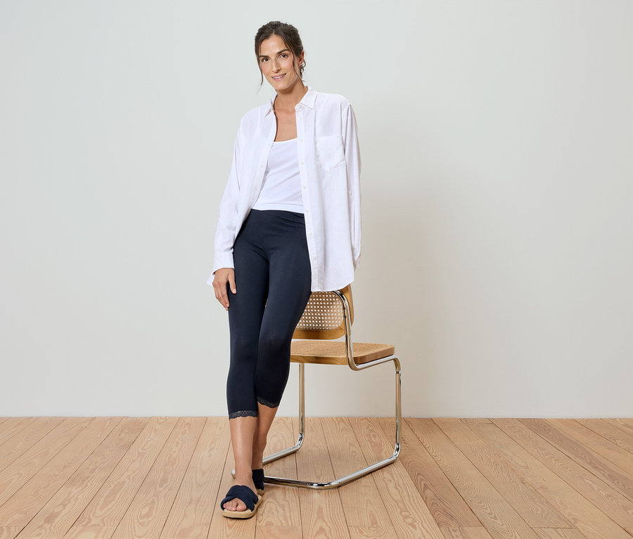 Une femme est appuyée sur une chaise, portant une chemise blanche, un débardeur blanc, un legging foncé et des sandales bleues.