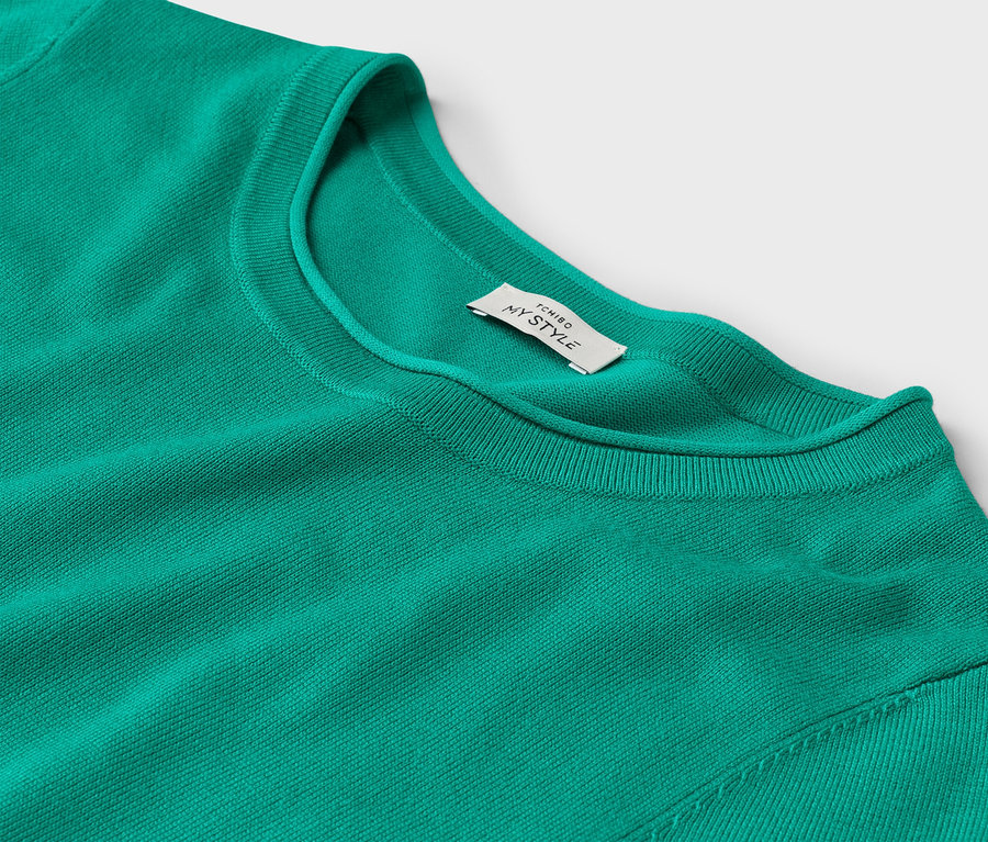 Gros plan d'un pull vert émeraude à col rond avec une étiquette indiquant « TCHIBO MY STYLE » sur un fond blanc.