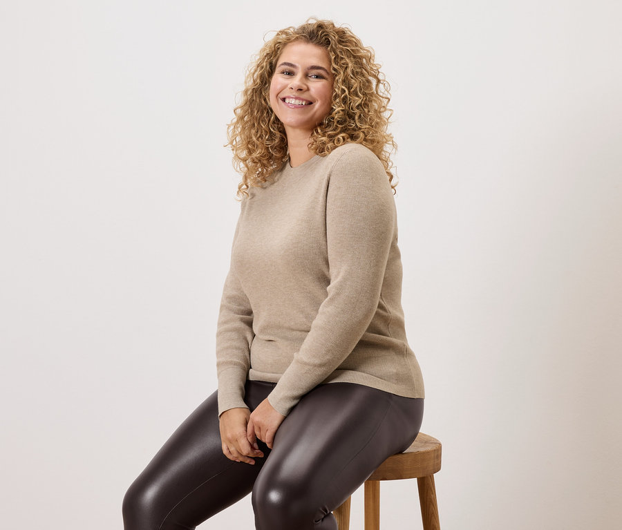 Une femme aux cheveux bouclés est assise sur un tabouret en bois, portant un pull beige et un legging foncé.