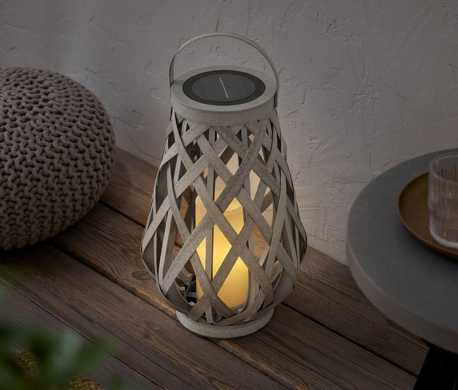 Une lanterne tressée avec un panneau solaire est posée à côté d'un pouf sur un plancher en bois.