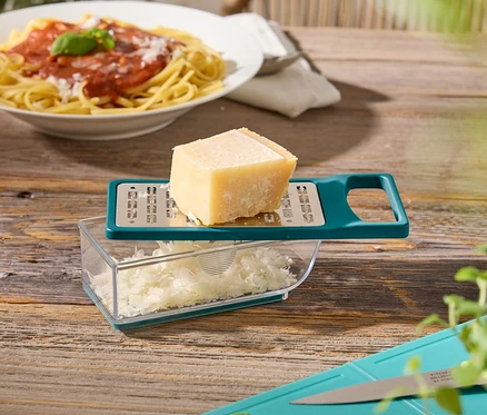 Sur une table en bois se trouvent une assiette de spaghettis, une râpe à fromage avec du fromage, une planche à découper avec du basilic et des courgettes.