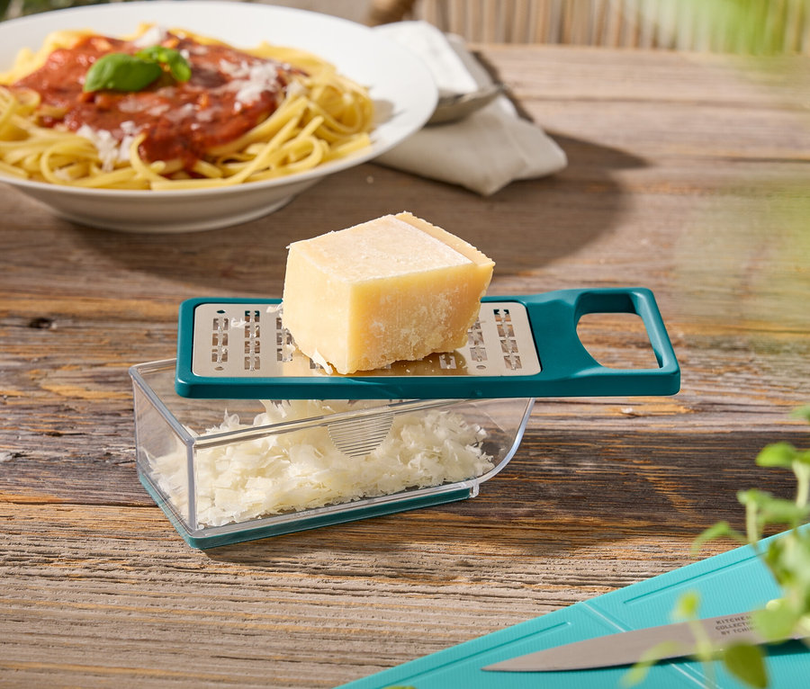 Sur une table en bois se trouvent une assiette de spaghettis, une râpe à fromage avec du fromage, une planche à découper avec du basilic et des courgettes.