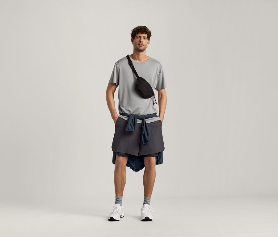 Un homme porte un t-shirt de sport gris clair chiné, un sac banane noir, un short et une veste nouée autour de la taille.