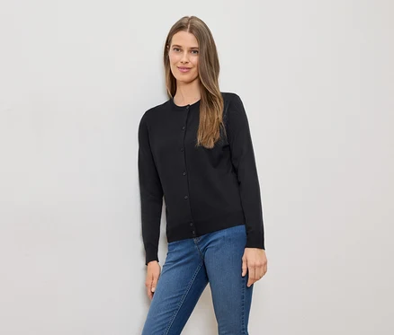 Une femme aux longs cheveux bruns, portant un cardigan noir et un jean bleu, est appuyée contre un mur blanc.