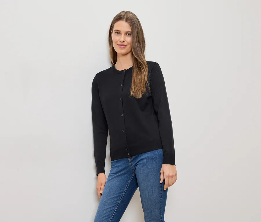 Une femme aux longs cheveux bruns, portant un cardigan noir et un jean bleu, est appuyée contre un mur blanc.