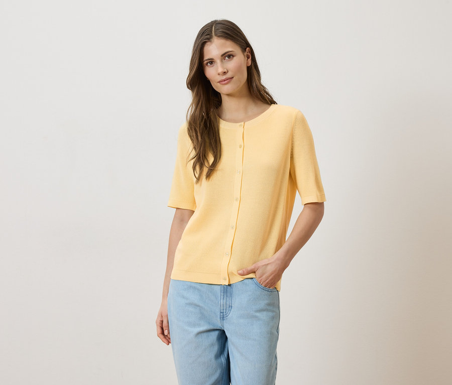 Une femme aux cheveux châtain clair porte un cardigan jaune clair à manches courtes boutonné et un jean bleu clair.