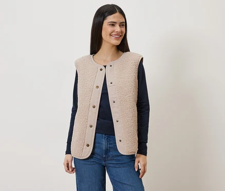Une femme aux longs cheveux bruns porte un gilet sans manches en fausse fourrure beige sur un t-shirt bleu marine à manches longues et un jean.