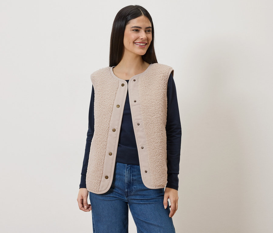 Une femme aux longs cheveux bruns porte un gilet sans manches en fausse fourrure beige sur un t-shirt bleu marine à manches longues et un jean.