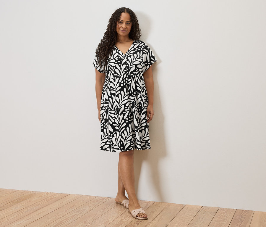 Femme en robe noire et blanche à motif avec encolure en V et sandales, debout devant un mur blanc.