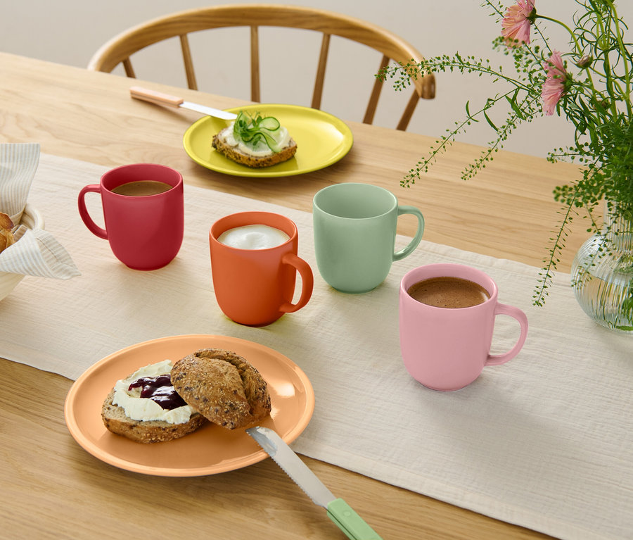 Table en bois avec chemin de table clair, quatre tasses colorées avec des boissons, deux assiettes avec des petits pains, un panier de pains et un vase de fleurs.