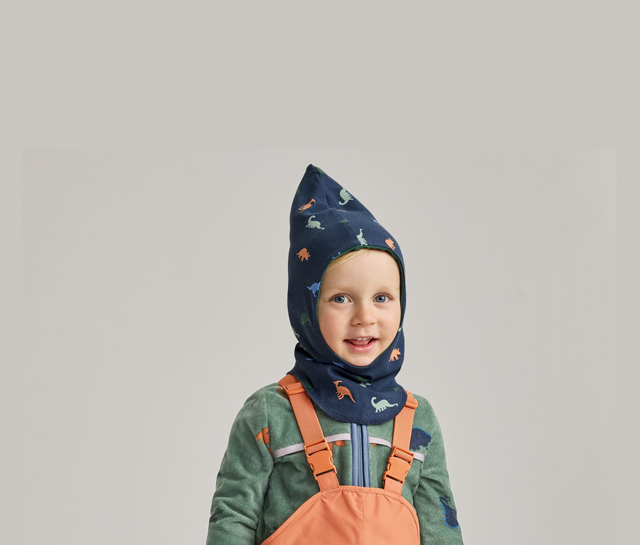 Un garçon souriant portant une cagoule bleue à motifs de dinosaures, un pull en polaire vert et une salopette orange sur fond gris.