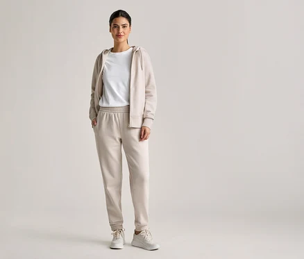 Une femme aux cheveux attachés porte un survêtement beige avec un sweat à capuche, un t-shirt et un pantalon de jogging, ainsi que des baskets blanches.