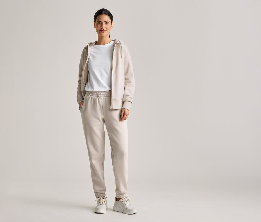 Une femme aux cheveux attachés porte un survêtement beige avec un sweat à capuche, un t-shirt et un pantalon de jogging, ainsi que des baskets blanches.