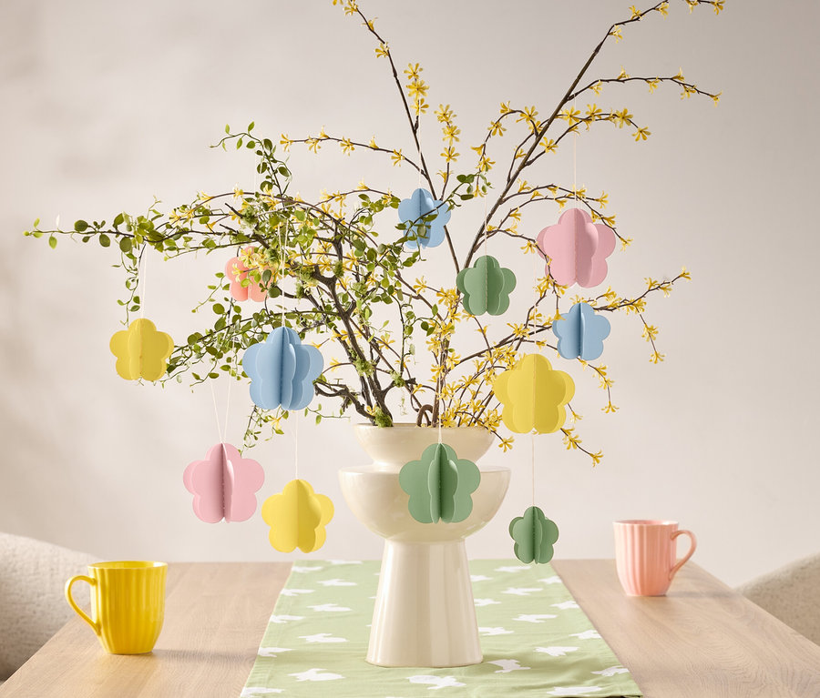 Des décorations florales sont suspendues à une branche dans un vase sur une table, à côté d'une tasse jaune et d'une tasse rose.