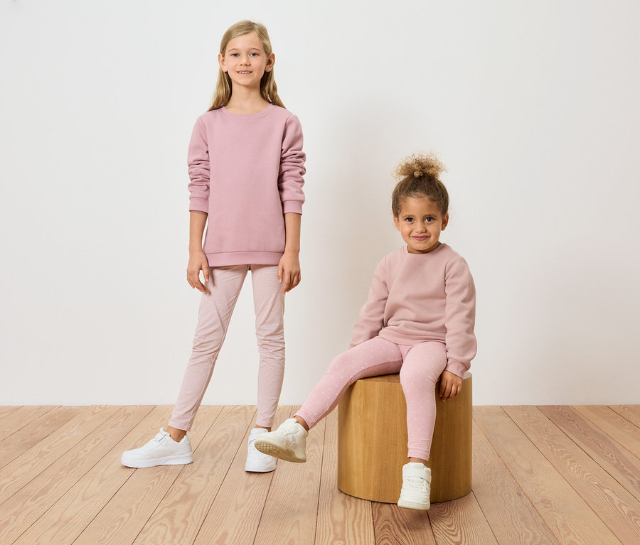 Deux filles, l'une debout et l'autre assise sur un tabouret, portant des sweat-shirts et des leggings roses assortis.