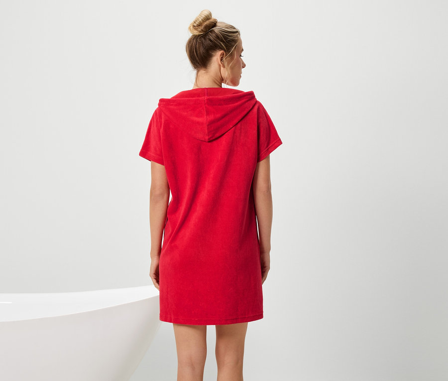 Une femme portant un chignon est vêtue d'une robe rouge à capuche et à manches courtes, faisant face à une baignoire.
