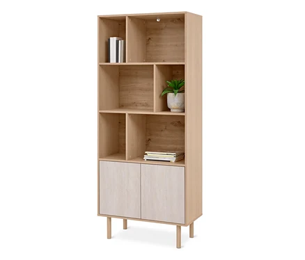 Étagère en bois clair avec des compartiments ouverts et deux portes d'armoire plus claires en bas. Elle contient des livres blancs, une plante verte dans un pot gris et des magazines. L'étagère repose sur quatre pieds courts.