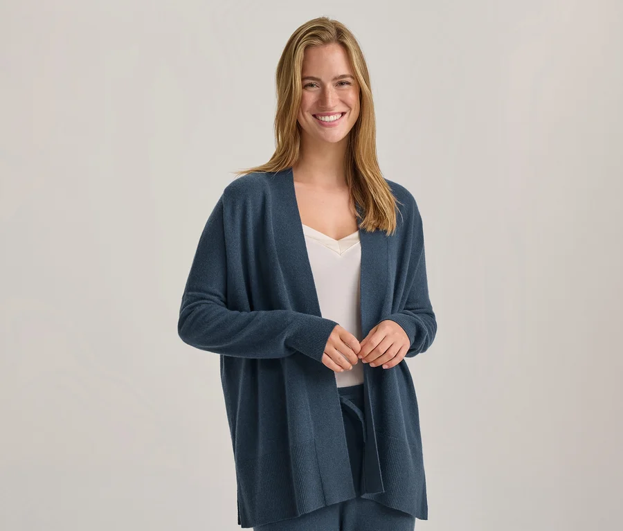 Femme souriante aux longs cheveux blonds portant un cardigan bleu ouvert sur un haut blanc et un pantalon bleu, les mains jointes.