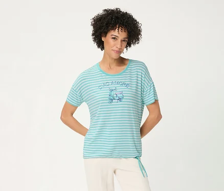 Femme souriante aux cheveux bouclés portant un T-shirt Olsen rayé turquoise et blanc avec un motif de scooter 'CIAO AMORE' et un pantalon clair, les mains derrière le dos.