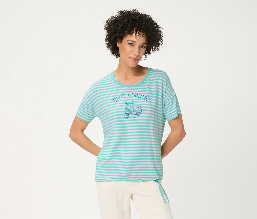 Femme souriante aux cheveux bouclés portant un T-shirt Olsen rayé turquoise et blanc avec un motif de scooter 'CIAO AMORE' et un pantalon clair, les mains derrière le dos.