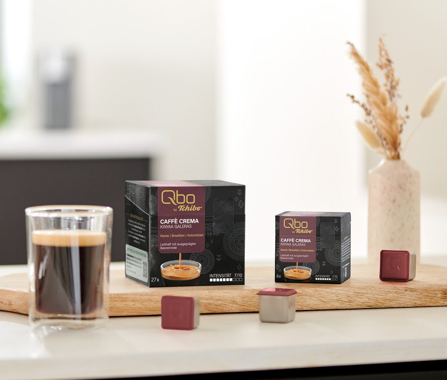 Un verre de café est posé à côté des paquets de capsules Qbo Caffè KINYAA GALERAS sur une planche en bois.