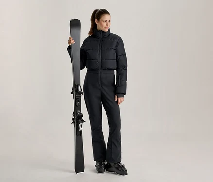 Femme portant une doudoune de ski courte noire et un pantalon de ski noir, tenant un ski noir verticalement et chaussée de bottes de ski noires.