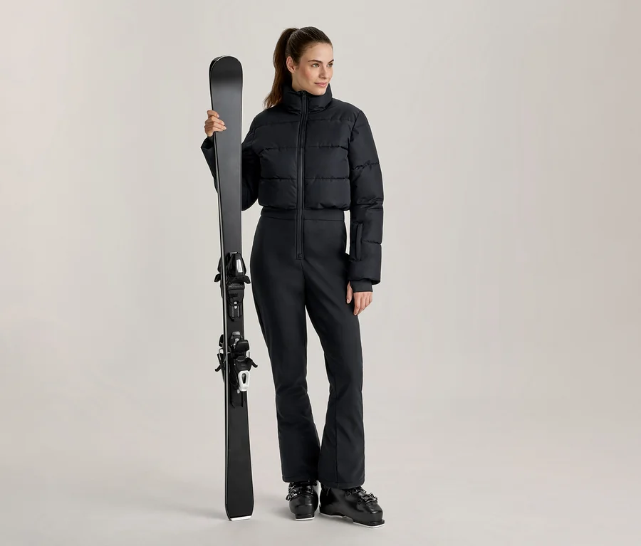 Femme portant une doudoune de ski courte noire et un pantalon de ski noir, tenant un ski noir verticalement et chaussée de bottes de ski noires.