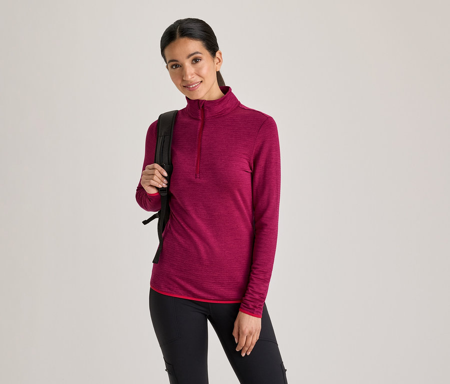 Femme portant un haut magenta à manches longues avec demi-fermeture éclair, un pantalon noir et un sac à dos noir, souriant à la caméra.
