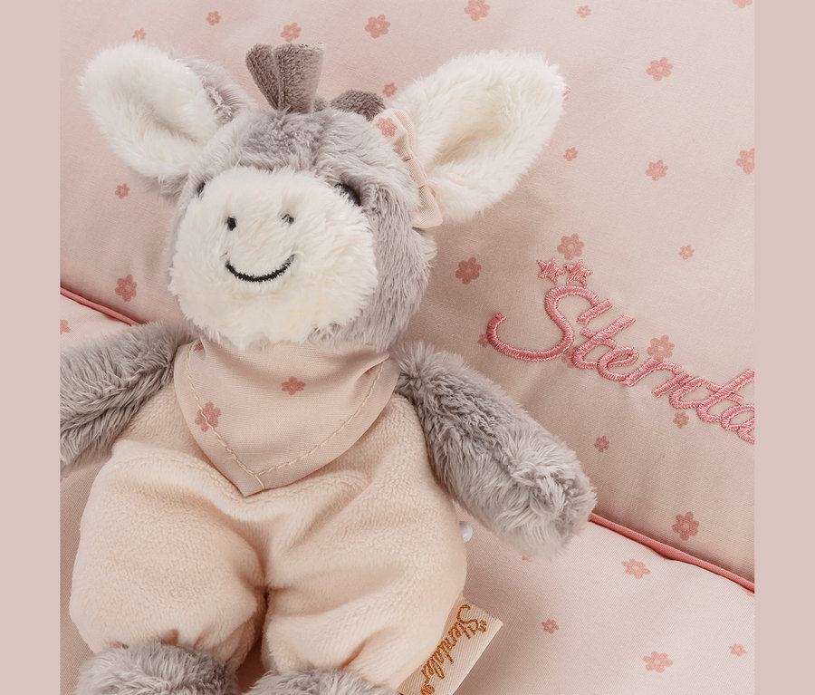 Un âne en peluche gris et blanc Sterntaler «Esel Emmi Girl» avec une salopette rose et un bandana, posé sur un tissu rose à motifs floraux.