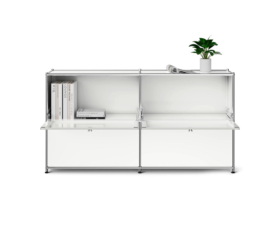 Un buffet modulaire blanc avec un cadre chromé. L'étagère supérieure gauche contient des livres, la droite est vide. Une plante et un journal roulé sont posés sur le côté droit. Les portes battantes inférieures sont légèrement ouvertes.