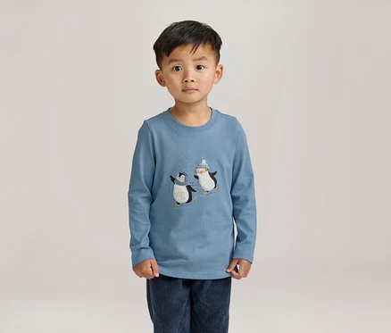 Un petit garçon se tient debout, portant un t-shirt bleu clair à manches longues avec deux pingouins patinant et un pantalon bleu foncé.
