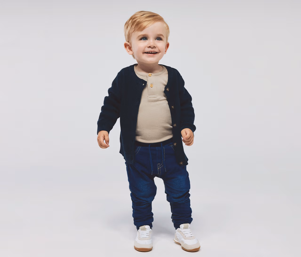 Un petit garçon souriant aux cheveux blonds et aux yeux bleus, portant un haut beige à manches longues, un cardigan en maille bleu marine, un jean bleu foncé et des baskets blanches et beiges.