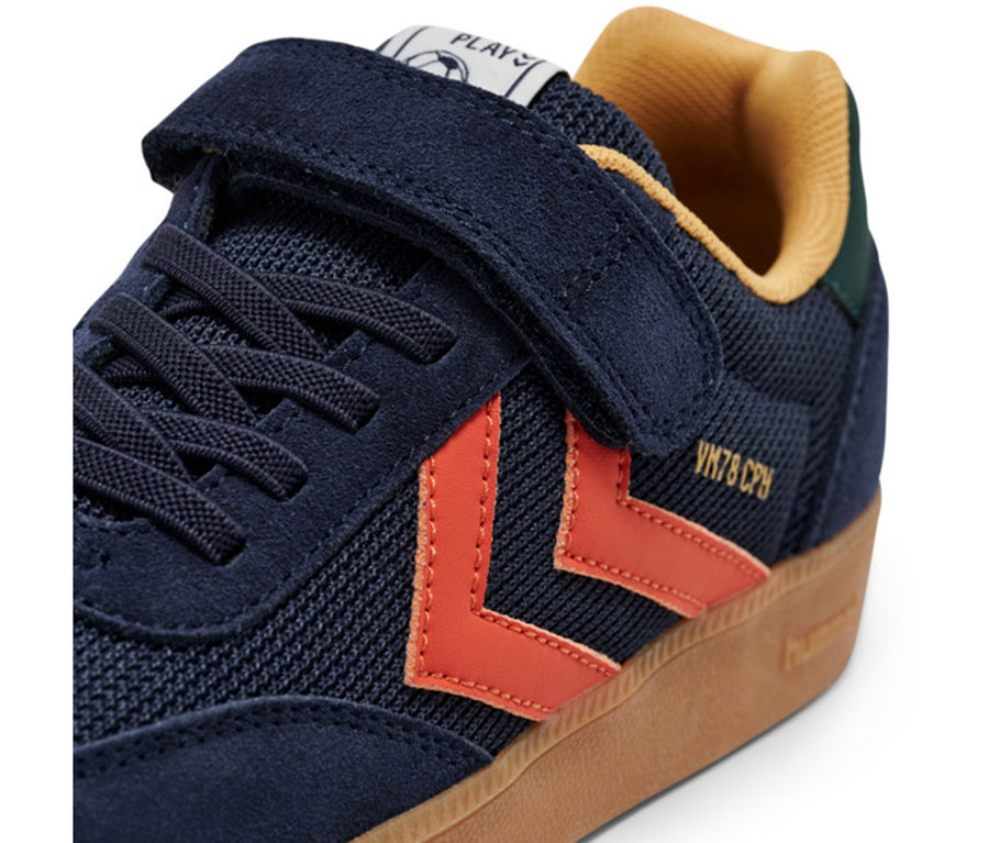 Gros plan sur une basket hummel® « VM78 CPH JR » multicolore, bleu foncé avec des chevrons orange, des lacets bleu foncé, une fermeture auto-agrippante et une semelle gomme marron clair.