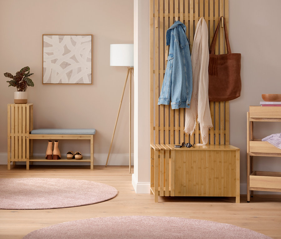 Un hall d'entrée lumineux avec un porte-manteau avec tablette où pendent des vestes. Au centre, un banc avec compartiment à chaussures et étagères en finition lamellé avec un coussin et des chaussures en dessous, à côté d'un lampadaire trépied en bois. À droite, une garde-robe avec banc, aspect lamellé, avec une veste en jean, une écharpe et un sac. À côté, une étagère sur pieds avec 3 compartiments. Deux tapis ronds roses sont posés au sol.
