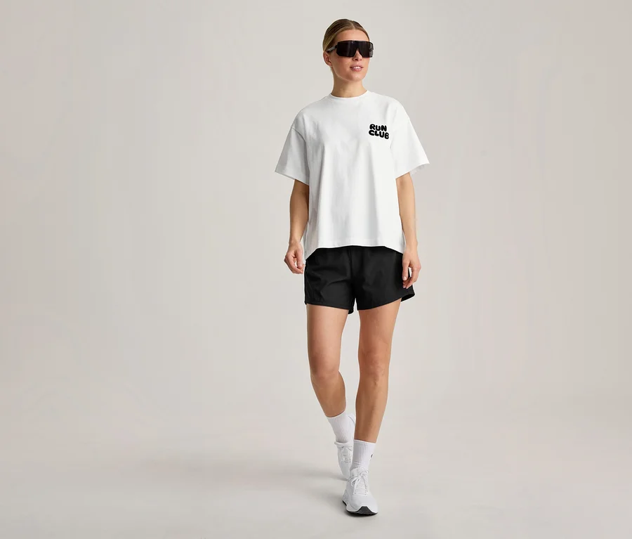 Une femme marche, vêtue d'un T-shirt de sport blanc avec l'inscription « RUN CLUB », d'un short noir, de chaussettes blanches, de baskets blanches et de lunettes de soleil noires.