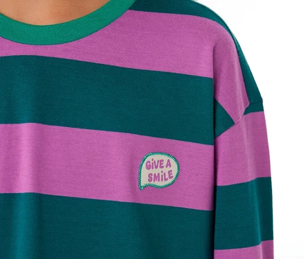 Gros plan d'un enfant portant un haut de pyjama SCHIESSER à rayures horizontales violettes et vert foncé, avec un col vert et un patch brodé "GIVE A SMILE" en forme de bulle.