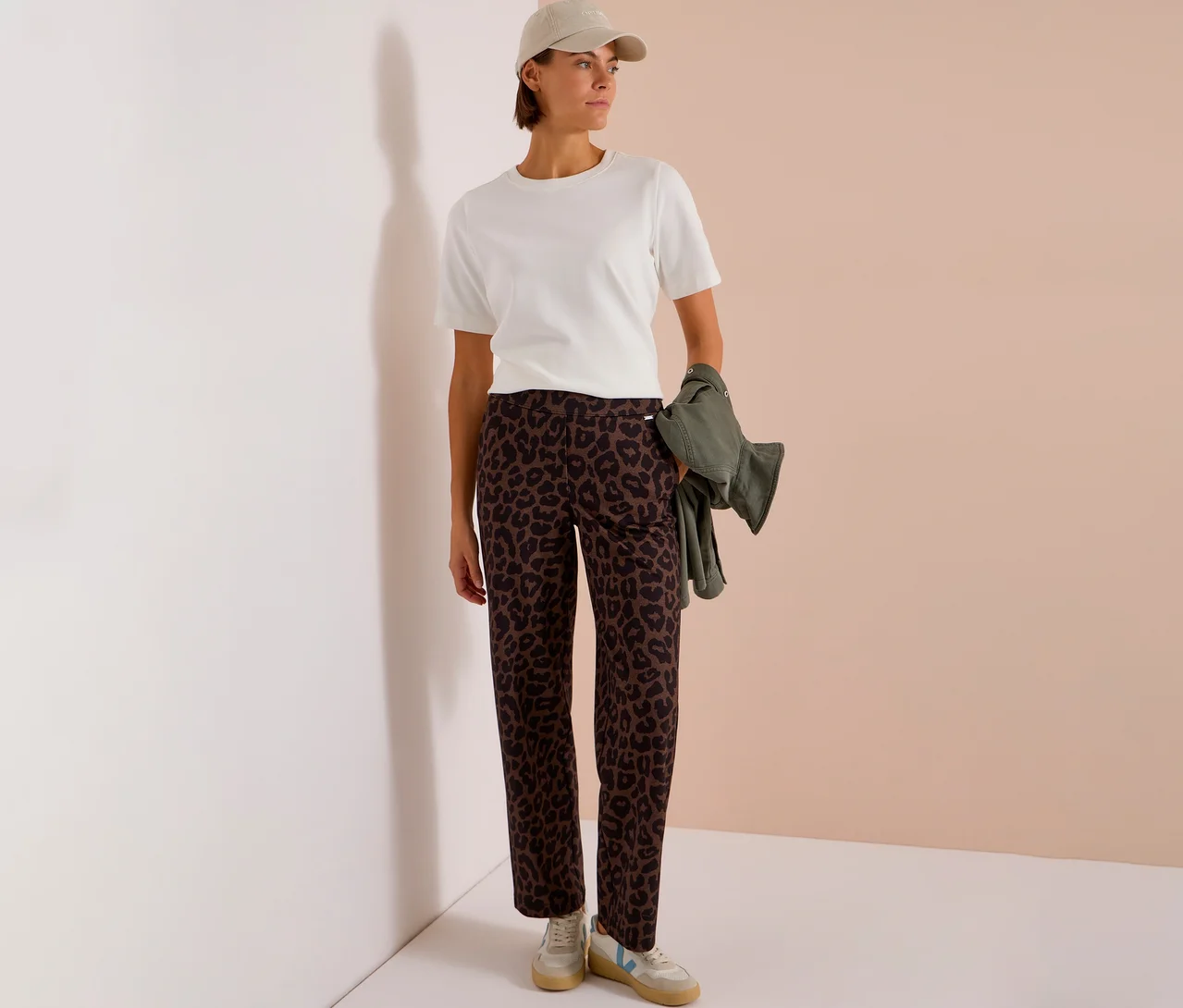 Femme portant un t-shirt blanc, une casquette beige et un pantalon en tissu CECIL imprimé sur toute la surface, tenant une veste vert olive.