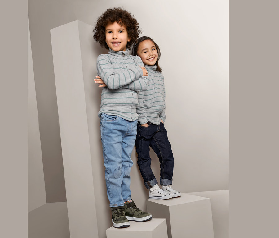 Deux enfants debout sur des blocs gris. Le garçon à gauche porte une jaquette en maille sportive, un jean et des chaussures vertes. La fille à droite porte une jaquette en maille sportive, un jean foncé et des chaussures blanches.