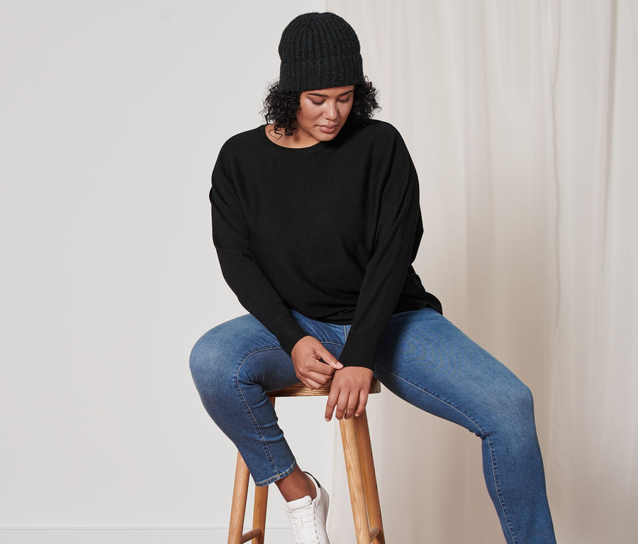 Femme assise sur un tabouret portant un pull en maille fine noir, un bonnet en maille anthracite avec de la laine et un jean slim en denim bleu moyen « Fit Emma ».