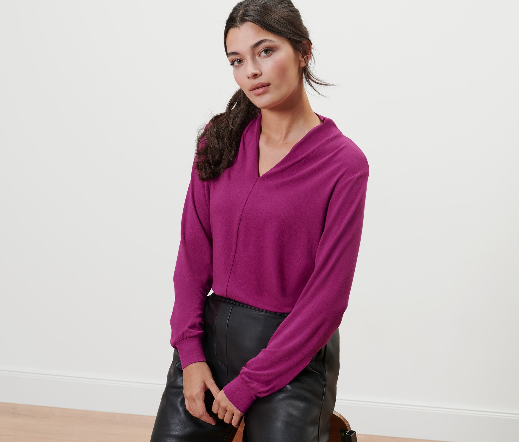 Une femme est assise sur un tabouret et porte un t-shirt à manches longues framboise avec un col en V.
