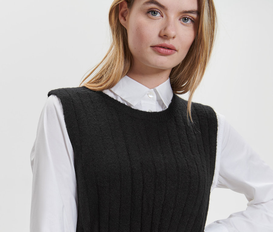 Une femme blonde porte une robe noire en tricot côtelée sur une chemise blanche.