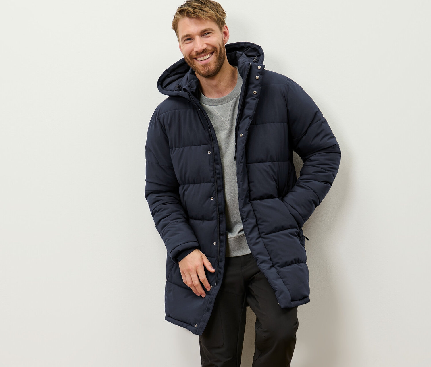 Un homme souriant s'appuie contre un mur et porte une parka matelassée avec capuche.