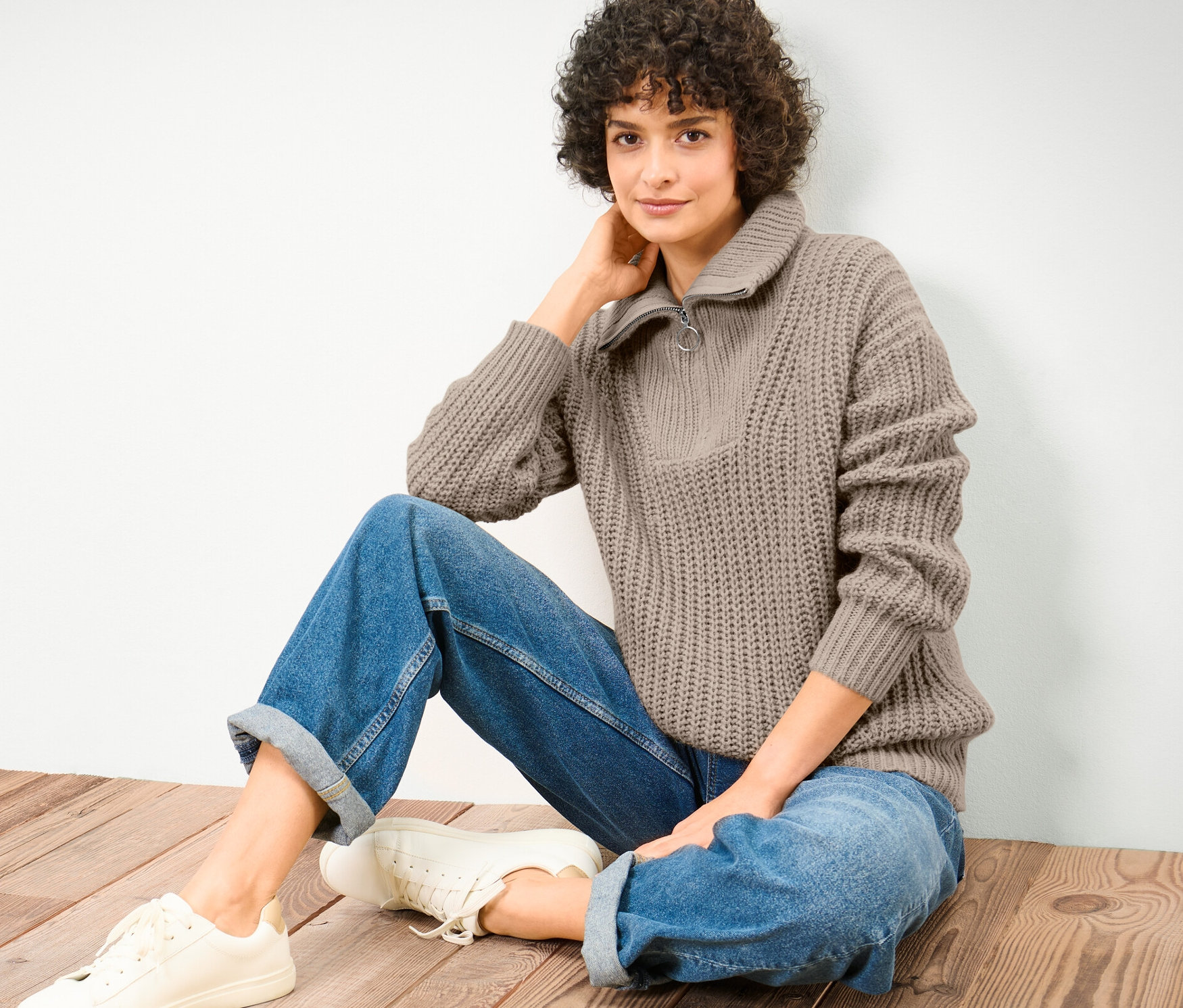 Femme assise sur un plancher en bois portant un pull camionneur taupe en grosse maille, un jean et des baskets.