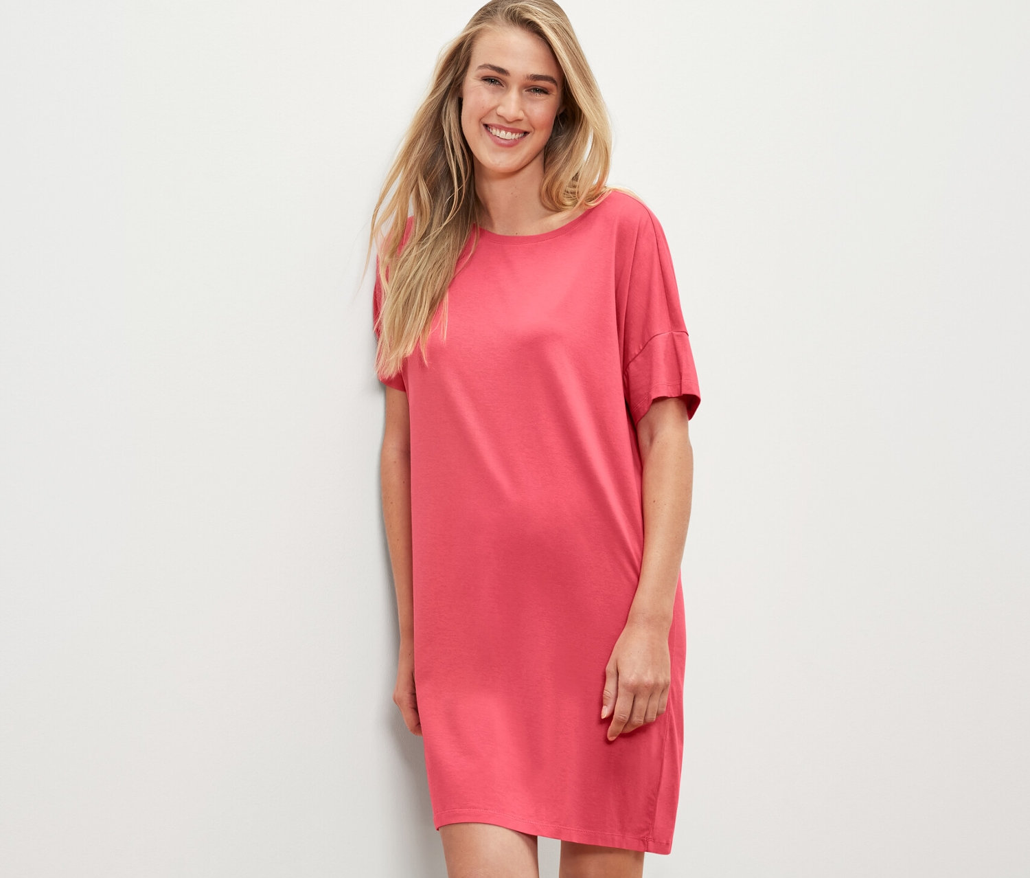 Femme blonde en chemise de nuit corail appuyée contre un mur blanc.