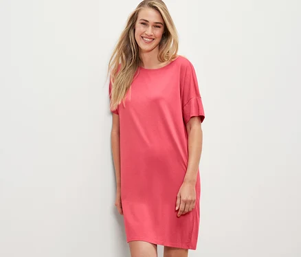 Femme blonde en chemise de nuit corail appuyée contre un mur blanc.